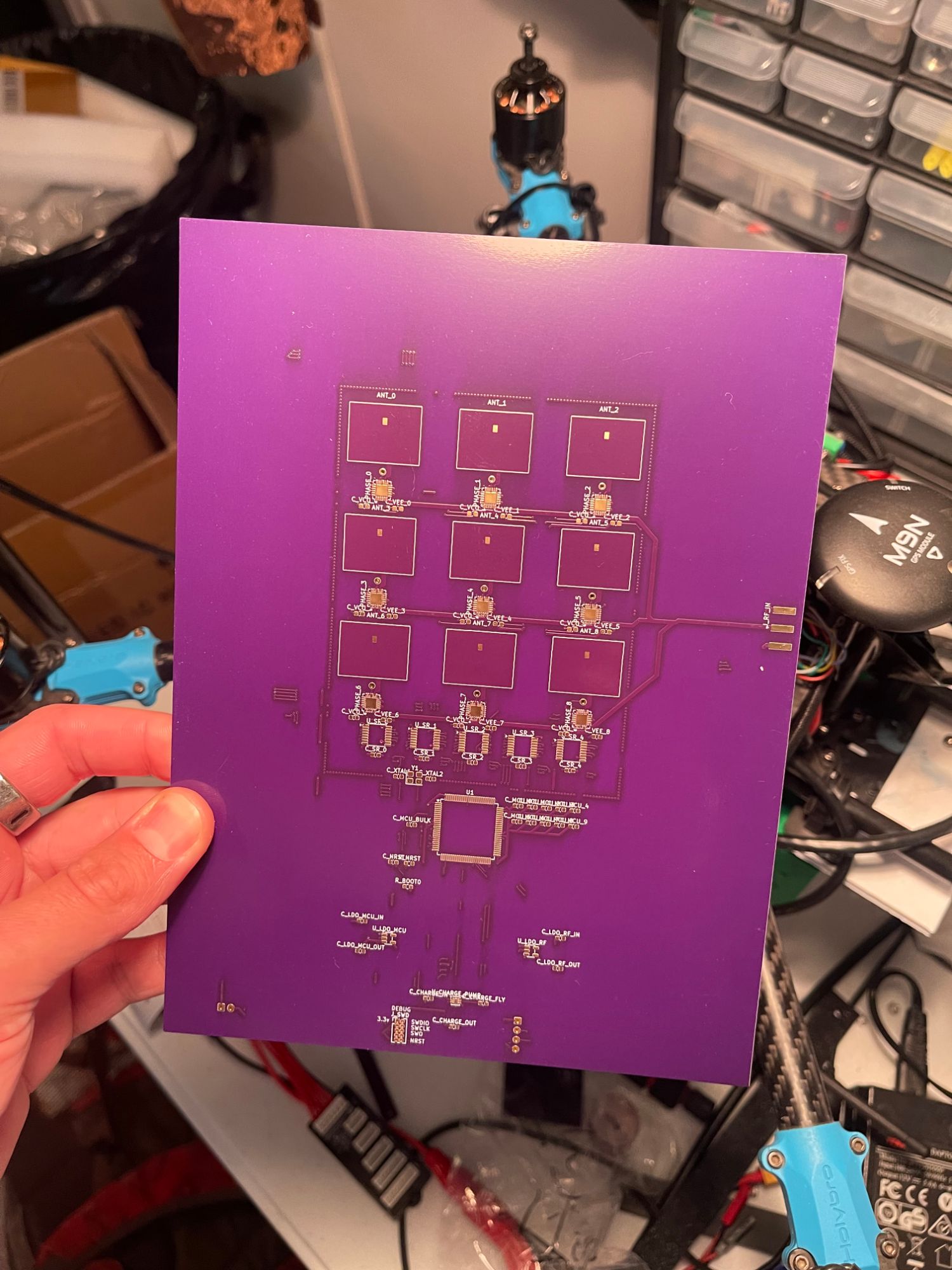 Fabricated Rev A 3x3 phased array PCB with purple soldermask, held in hand over an electronics workbench